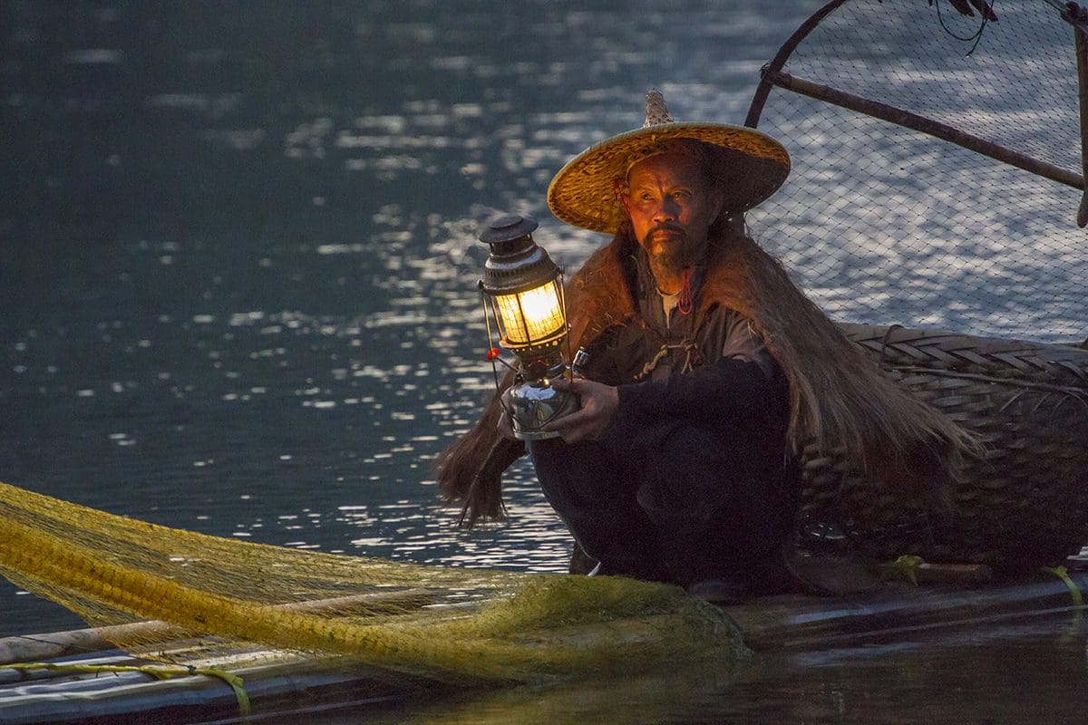 A man in a hat sits on a boat holding a lantern.