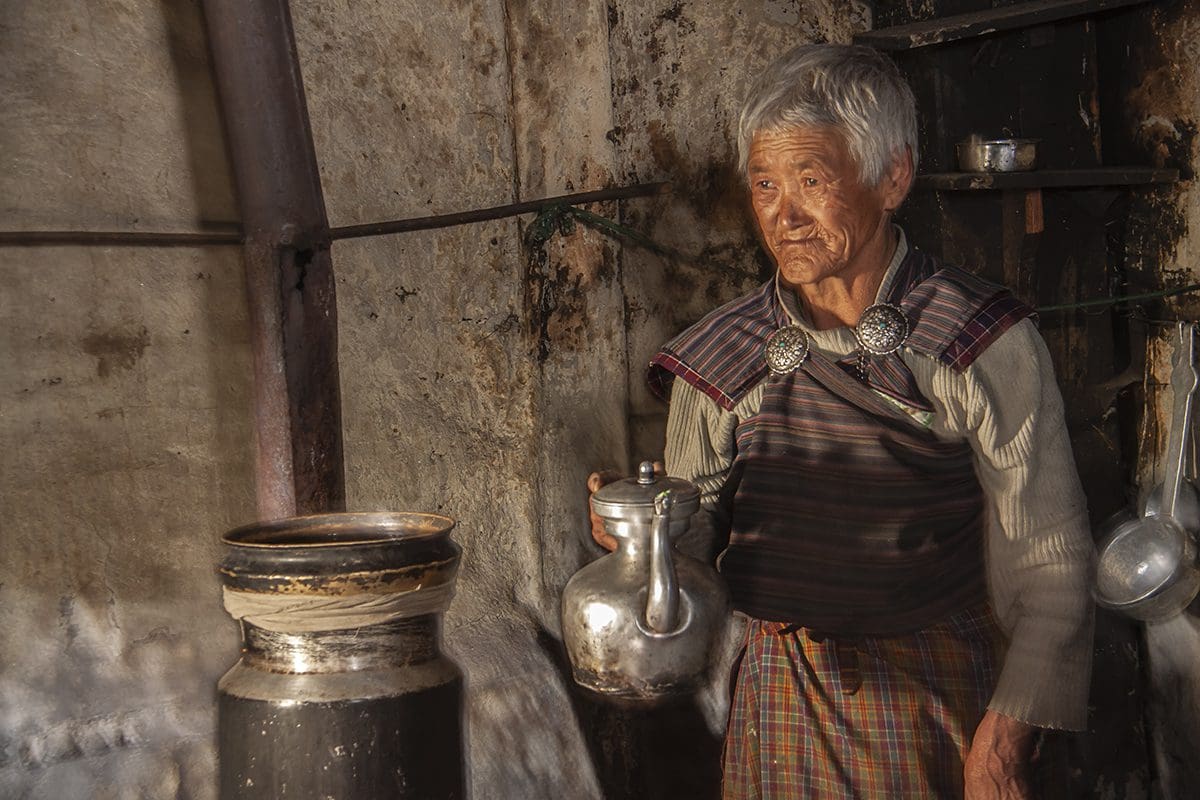 An old woman standing in a kitchen with a pot.