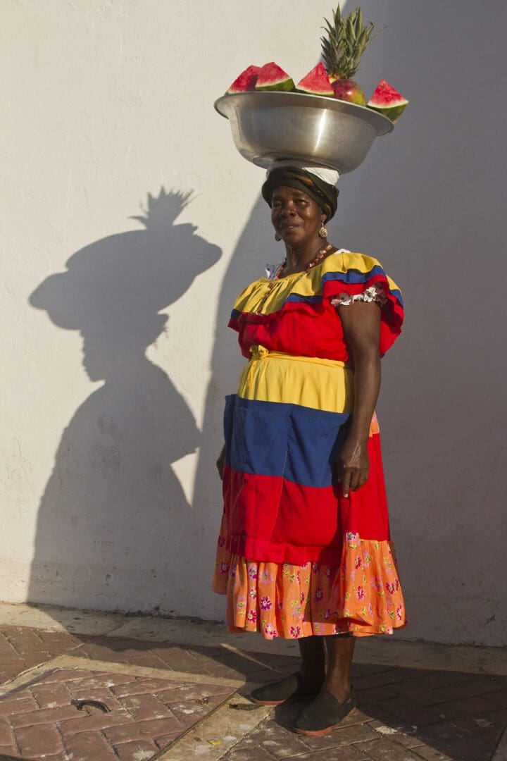 A woman in a colorful dress with a basket of fruit on her head.