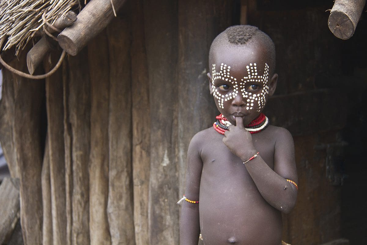 A young boy with a painted face standing in front of a hut.