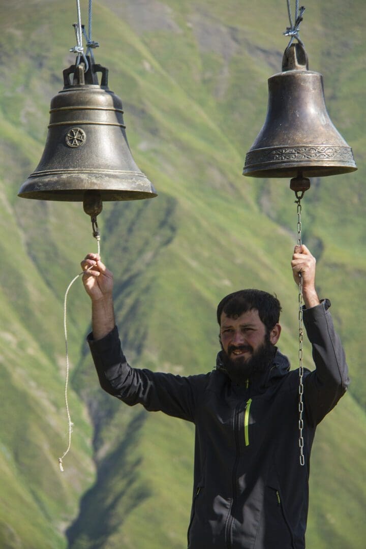 A man holding two bells in the mountains.