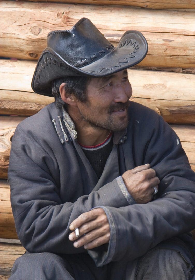 A man in a cowboy hat sitting next to a log cabin.