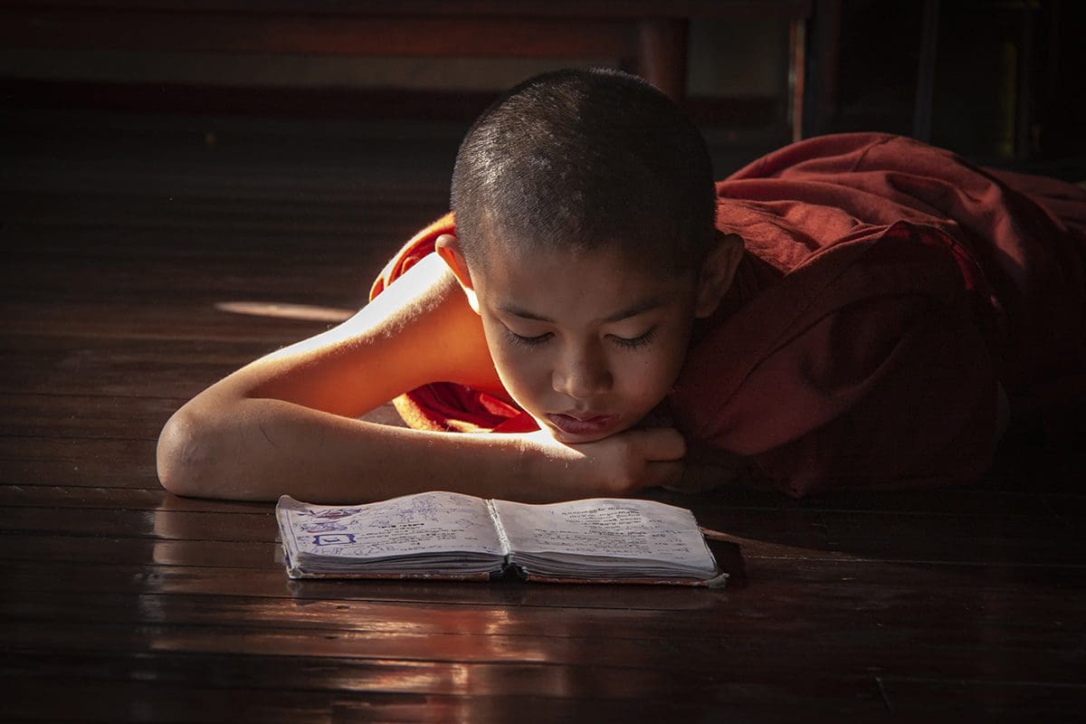 A young monk laying on the floor reading a book.