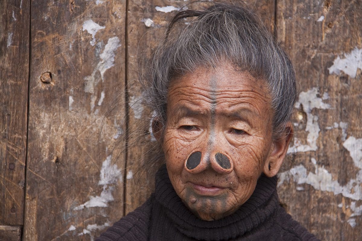 An old woman with a nose piercing in front of a wooden wall.