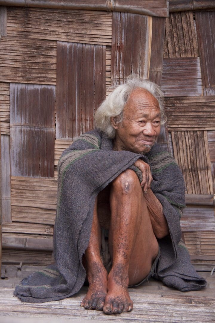 An old man sitting in front of a wooden hut.