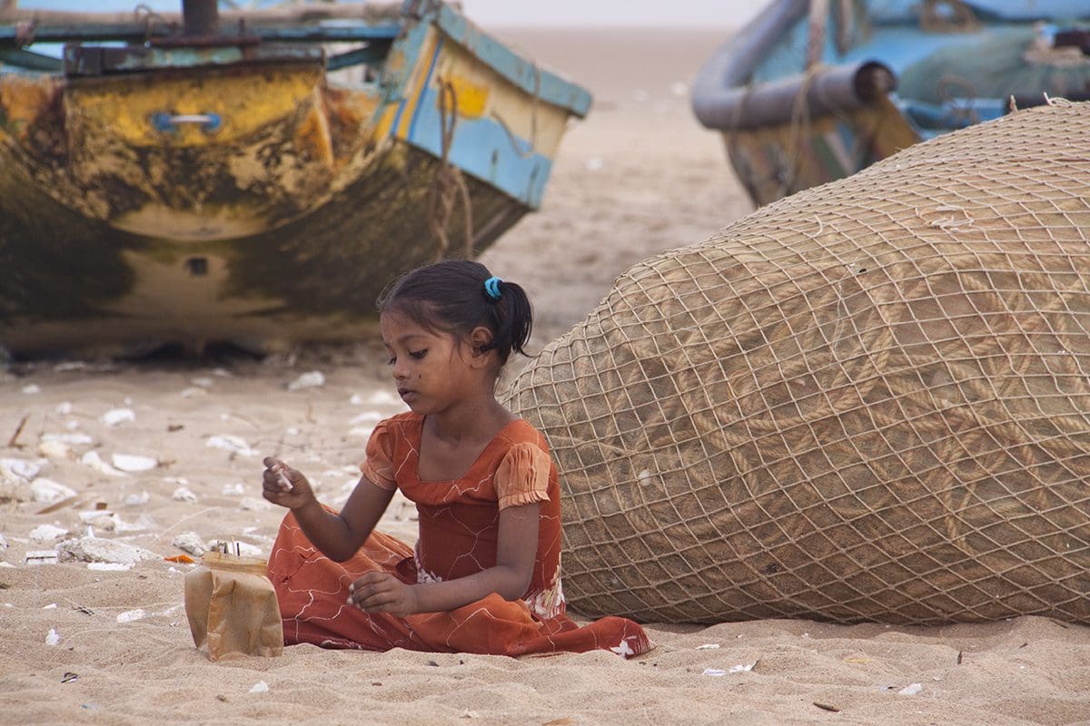 A girl sits on the sand next to a fishing boat.