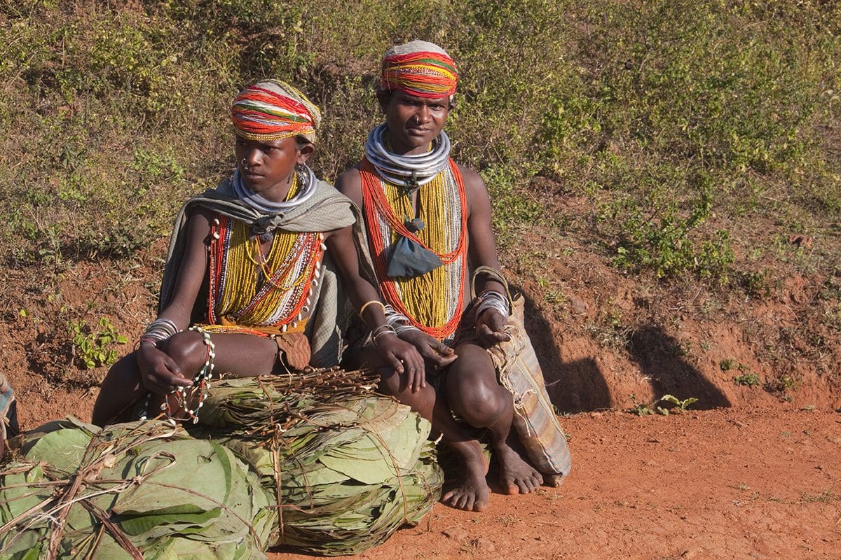 Two women sitting on a dirt road with bundles of leaves.