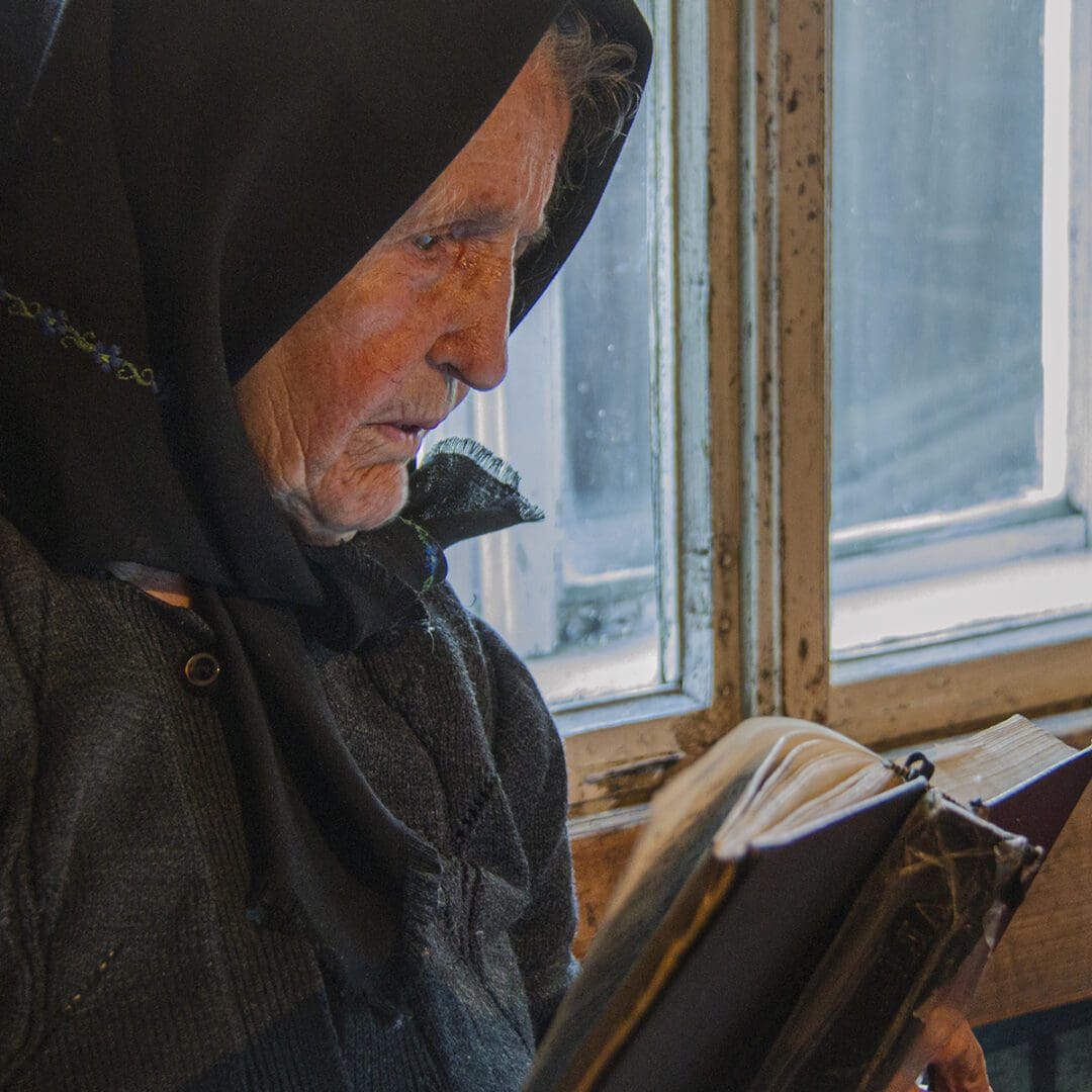 An old woman reading a book in front of a window.