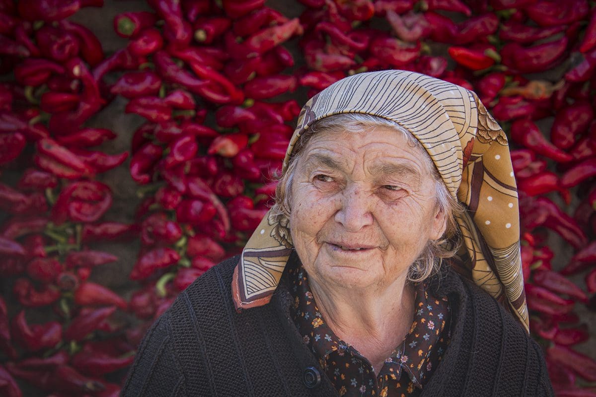 An old woman in front of a wall of red peppers.