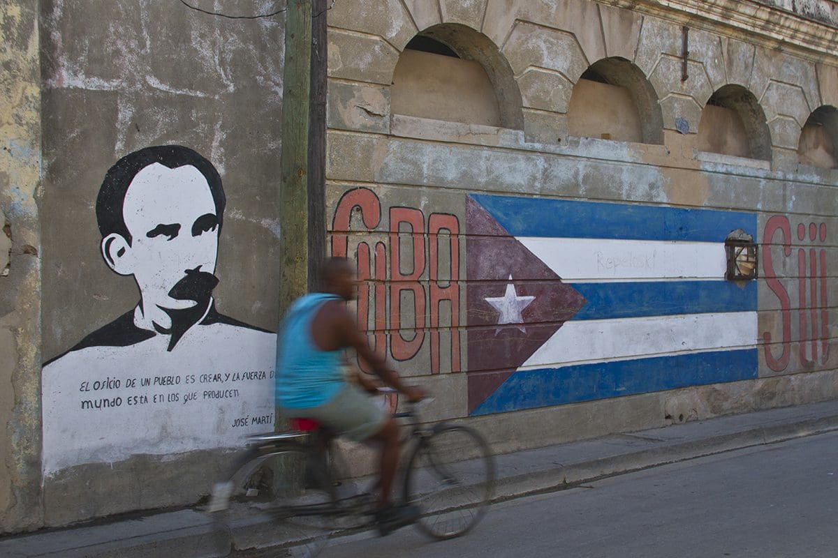 A man riding a bicycle past a mural of a cuban flag.