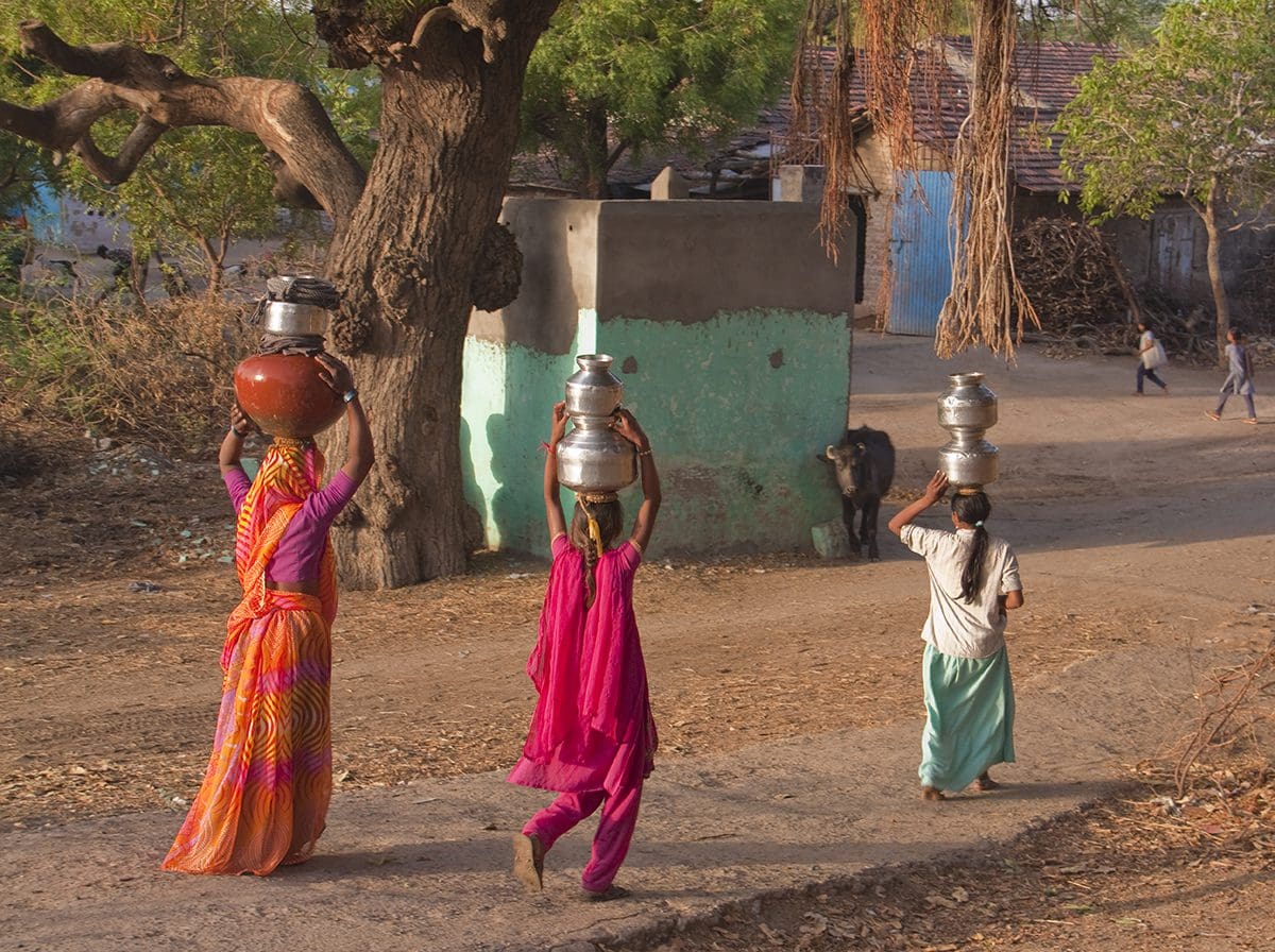 Three women carrying water jugs down a dirt road.