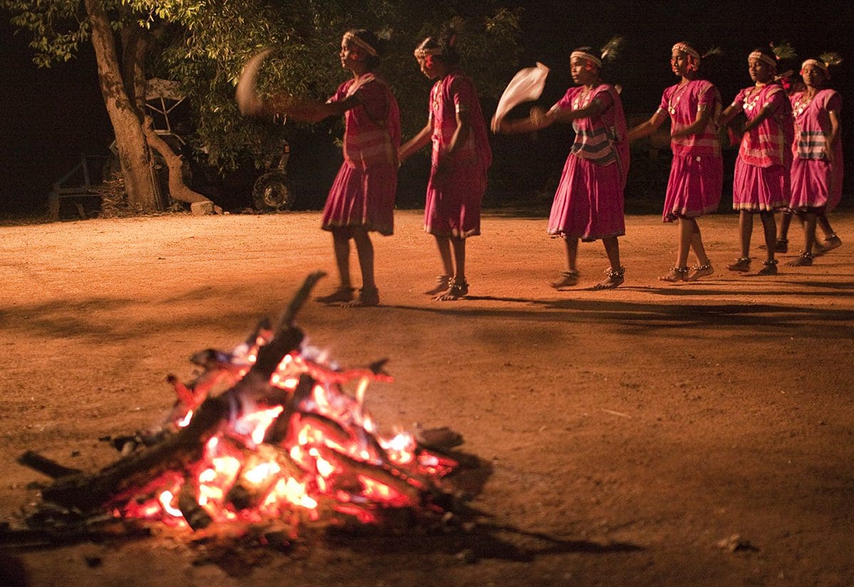 A group of women dance around a campfire at night.