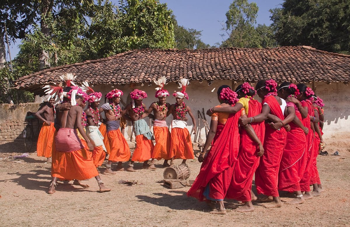 A group of women dressed in red and orange in front of a hut.
