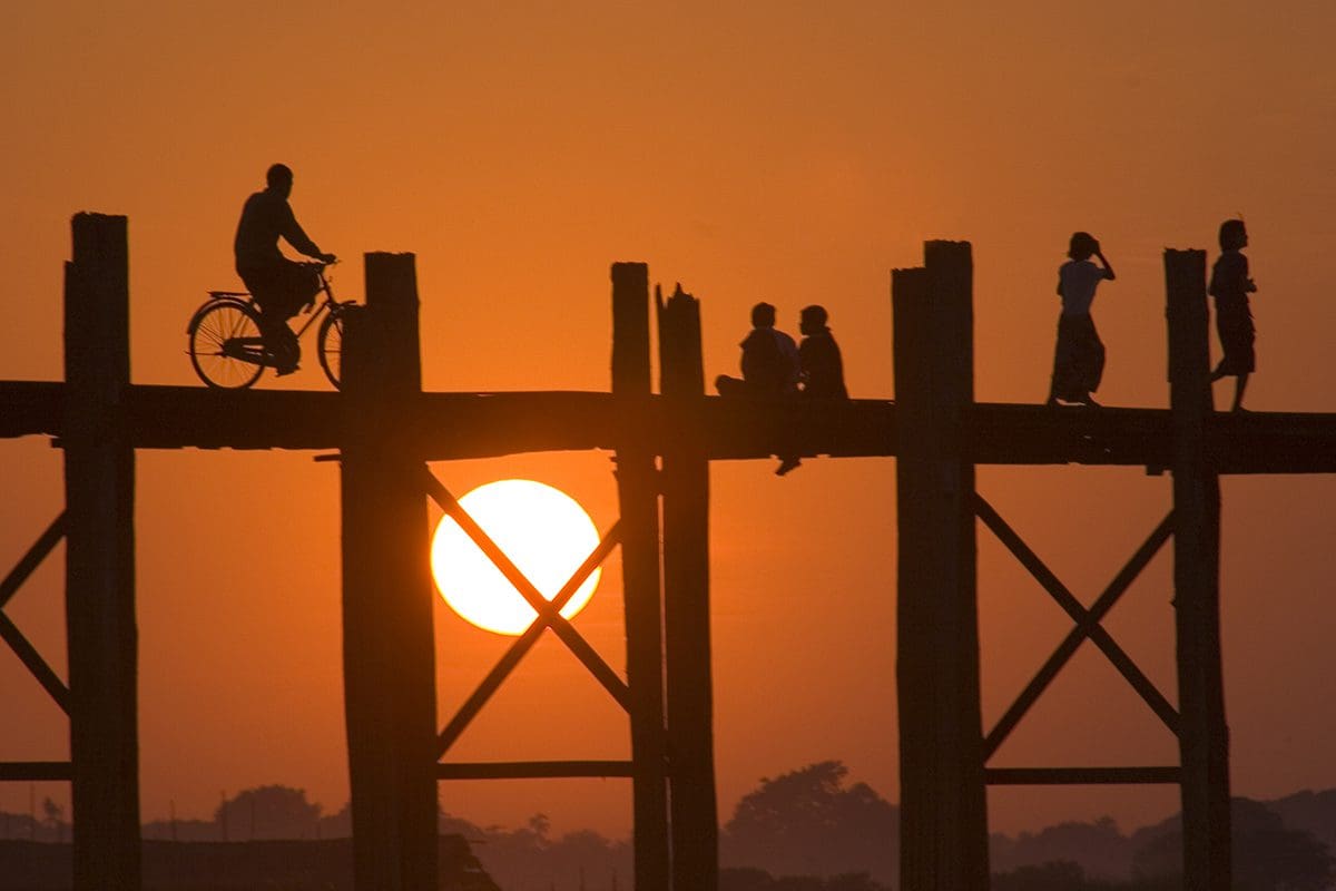 People riding bikes on a wooden bridge at sunset in myanmar.