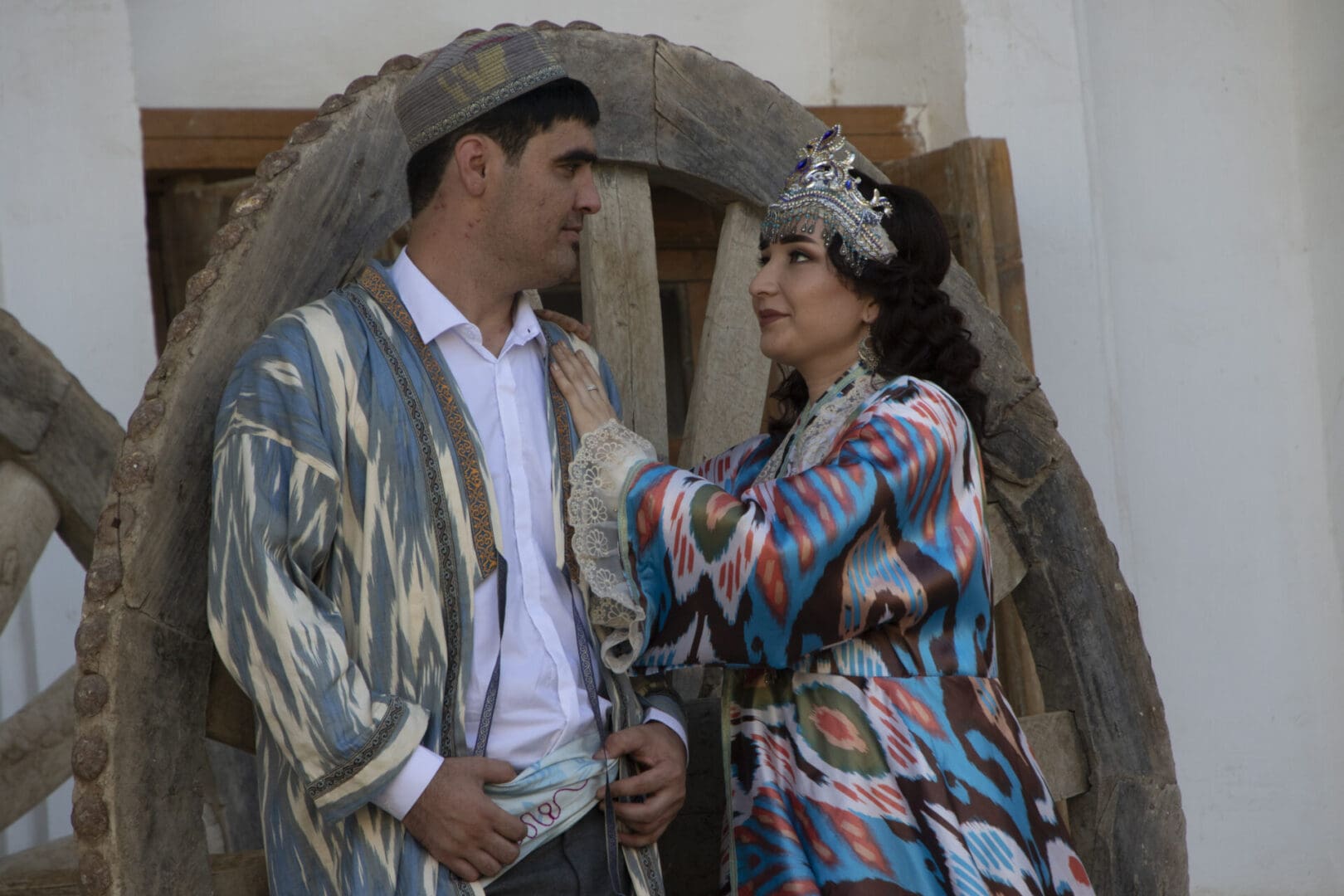 A man and a woman dressed in traditional clothing standing next to a wheel.